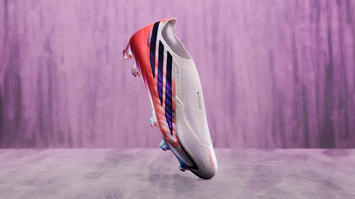 Chaussure de football adidas blanche à trois bandes violettes en diagonale, sans lacets, posée en équilibre sur la pointe devant un fond violet