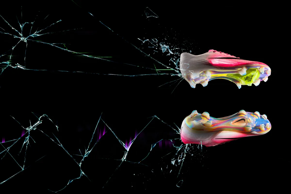 Deux chaussures de football Nike roses et blanches vues de dessous sur fond noir, l’une avec une unité Air Zoom jaune, l’autre avec une semelle translucide, devant une vitre brisée