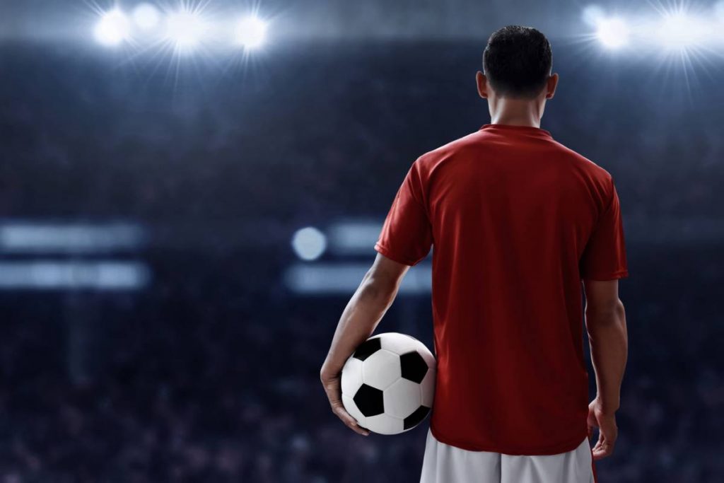 Joueur de dos en maillot rouge tenant un ballon dans un stade éclairé