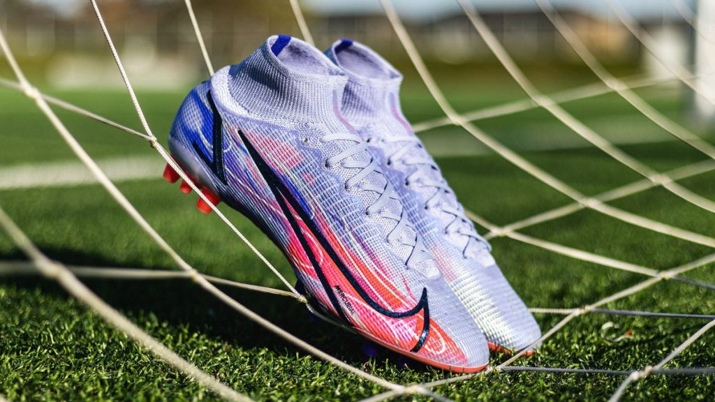 Chaussure de football Nike Mercurial blanche et rose posée contre un filet sur la pelouse