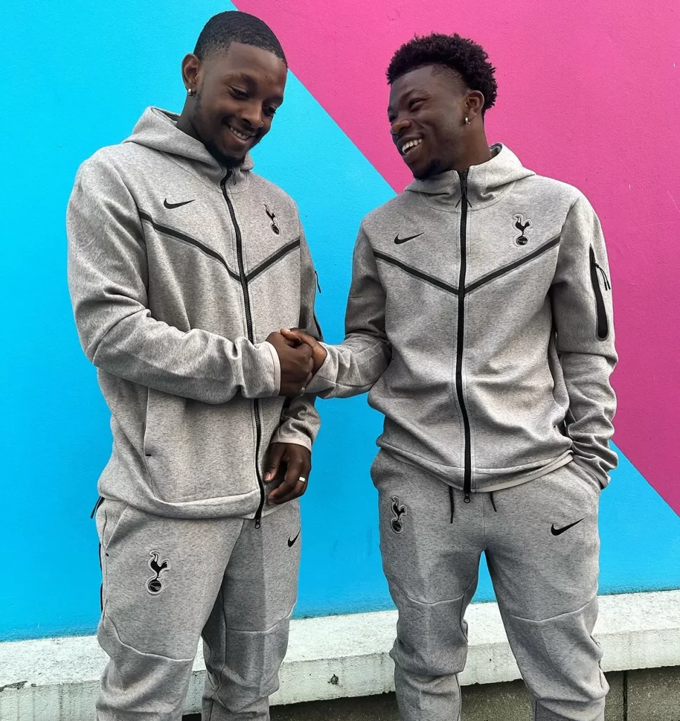 Deux hommes en survêtements Nike gris se serrant la main devant un fond bleu et rose