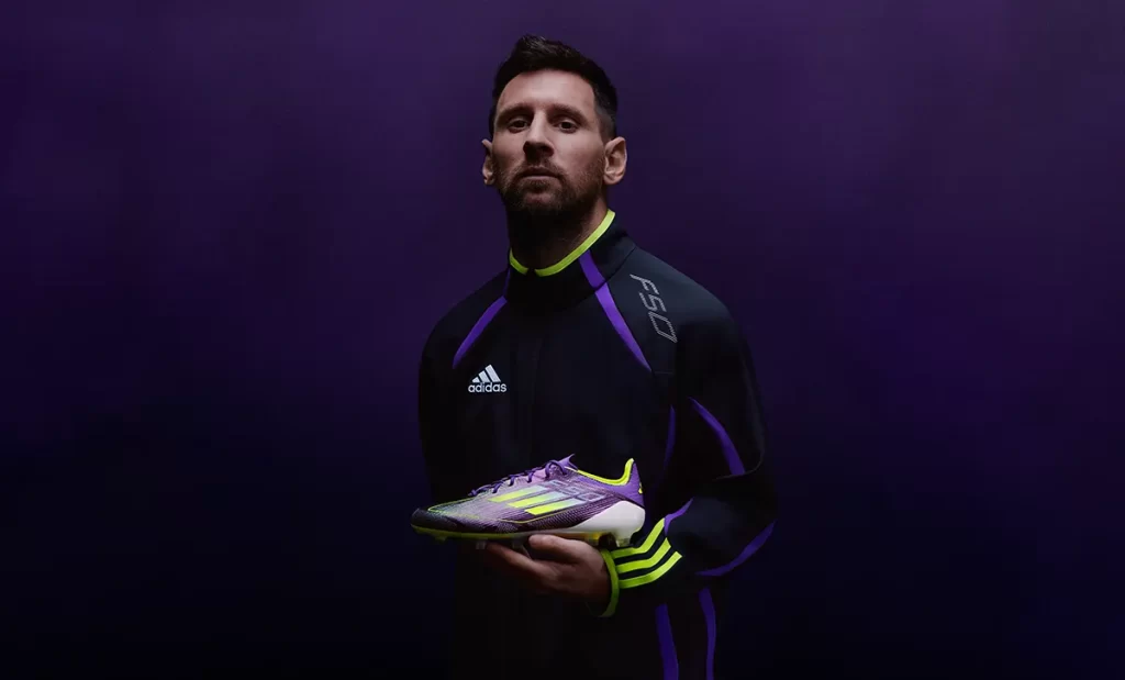 Joueur en veste noire tenant une chaussure de football adidas F50 blanche, sur fond violet